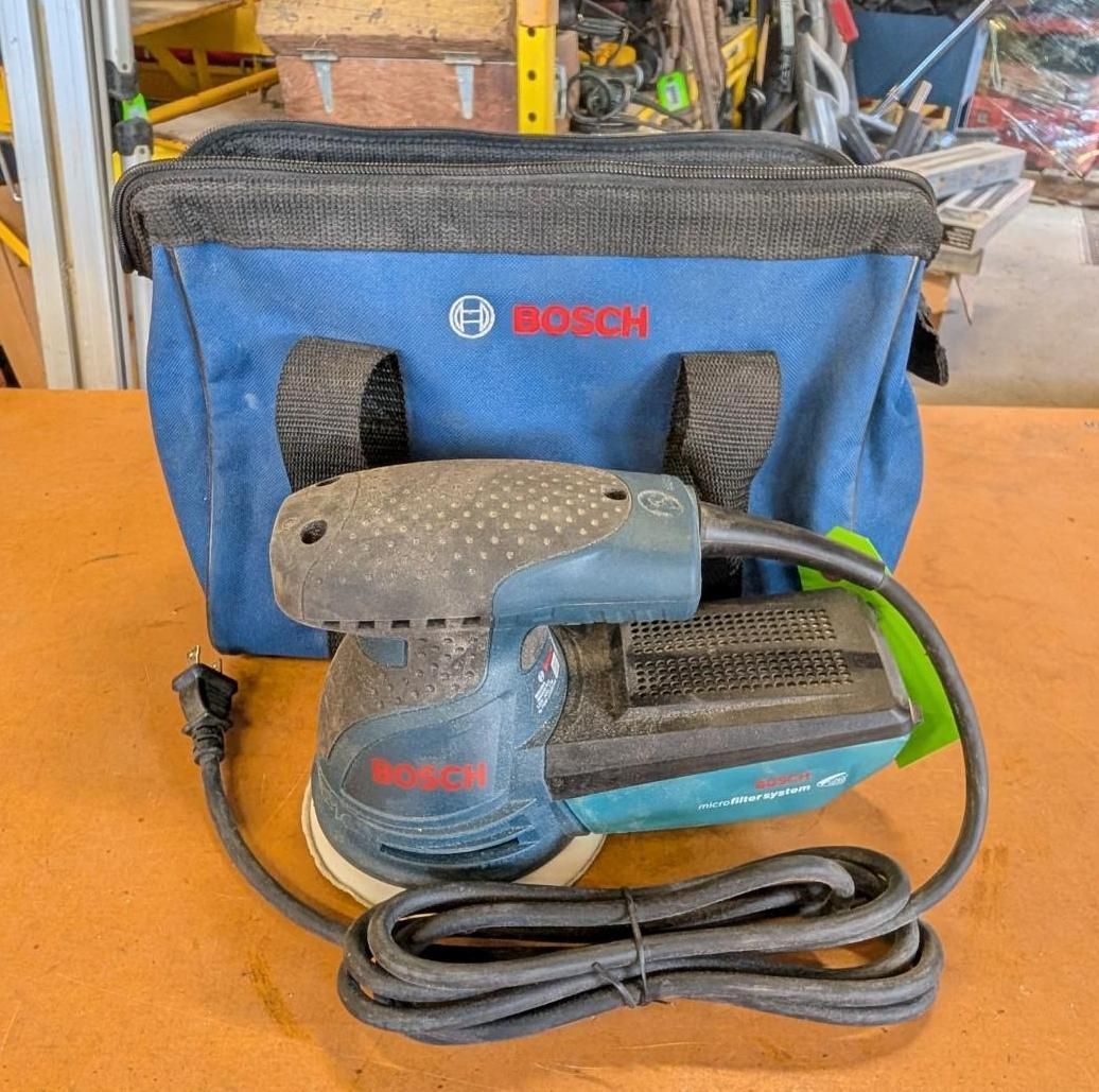 Blue Bosch orbital sander with cord in front of a blue Bosch tool bag in a workshop.