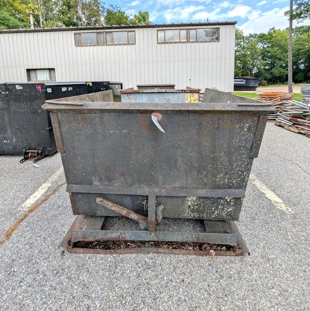 Large metal dumpster on a pallet in a gravel lot beside a white building.