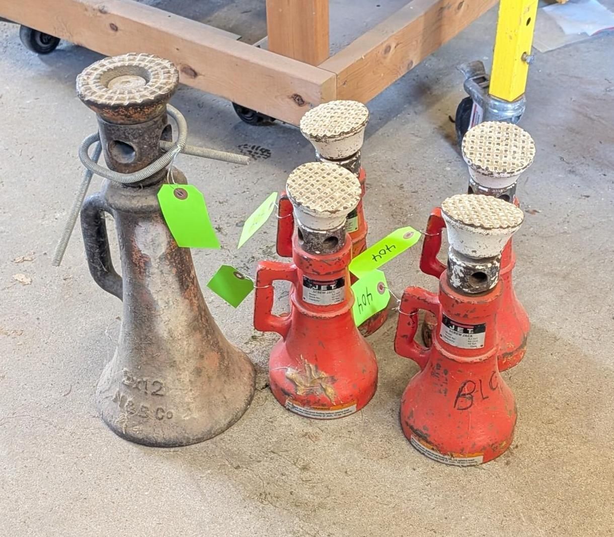 Three vintage metal bottle jacks on a concrete floor, one silver and two red, with bright green tags.