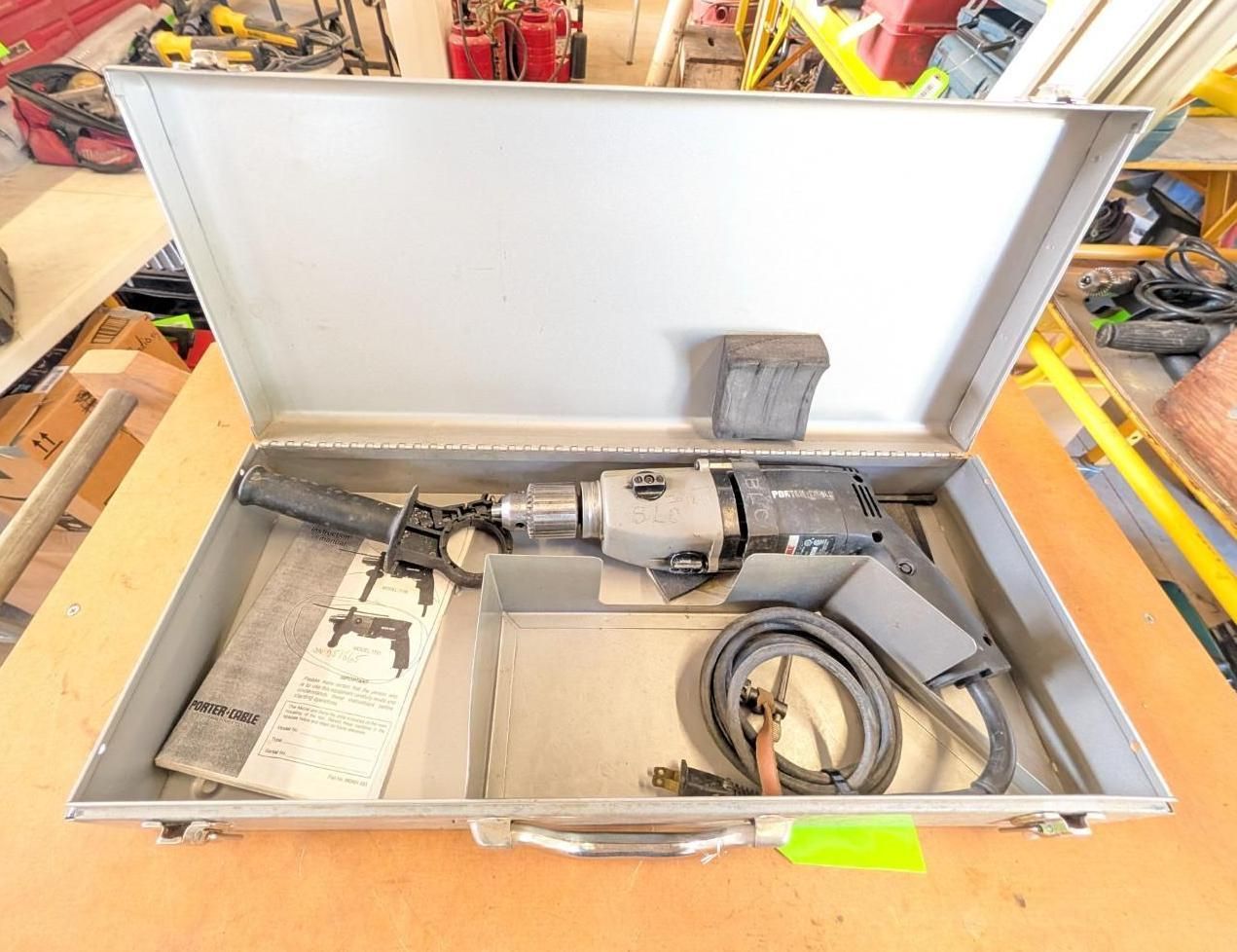Open metal case with a gray power drill, bit set, cable, and accessories on a workbench