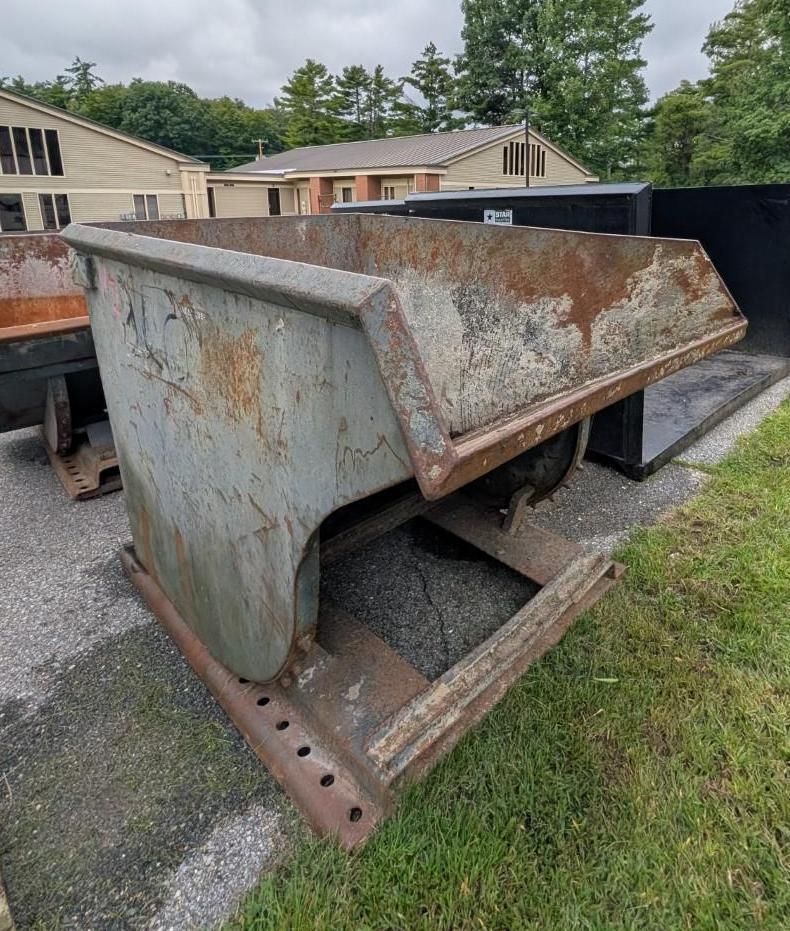 metal dumpster in a gravel lot near buildings and grass, viewed from the side.