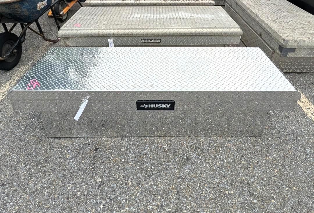 A silver diamond-plate truck box with a black label, set on a gray paved surface.