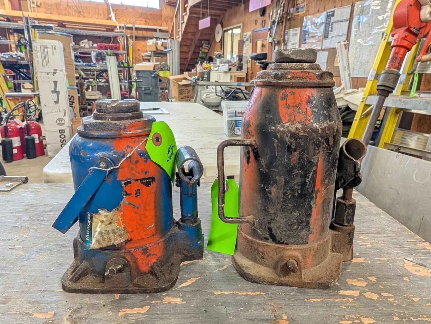 Two metal jacks on a workshop floor, one blue-orange and one black-orange, with tools in the background