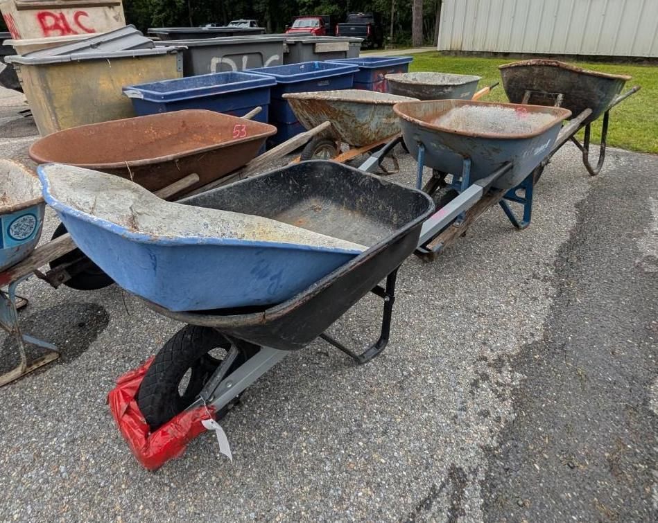 Several wheelbarrows lined up outdoors on pavement, one with a red bag in front