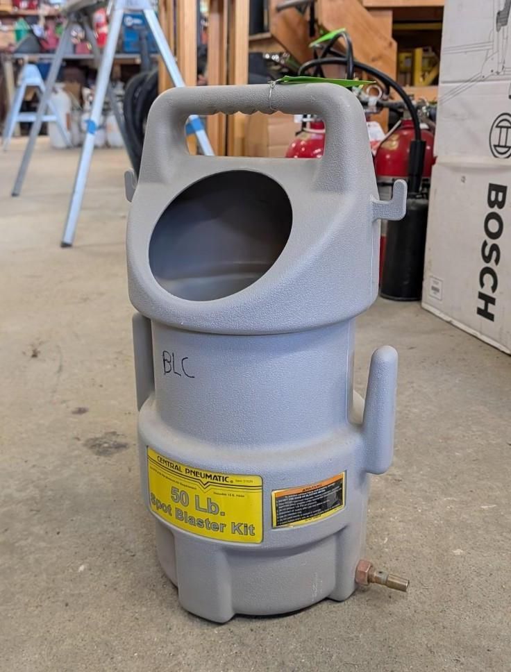 Gray industrial vacuum canister with handle and open top, standing in a workshop/garage.