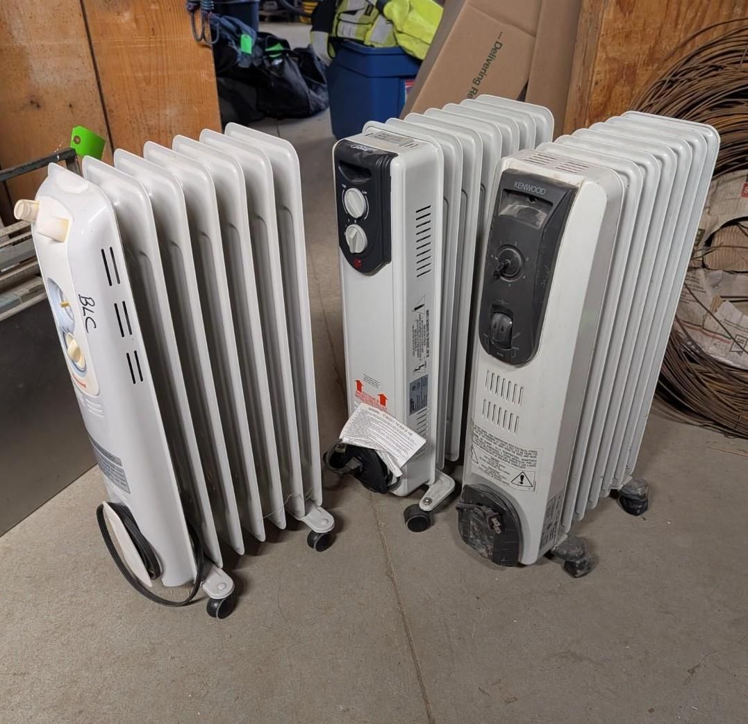 Three white electric radiators on casters lined up indoors, with cords visible on the floor