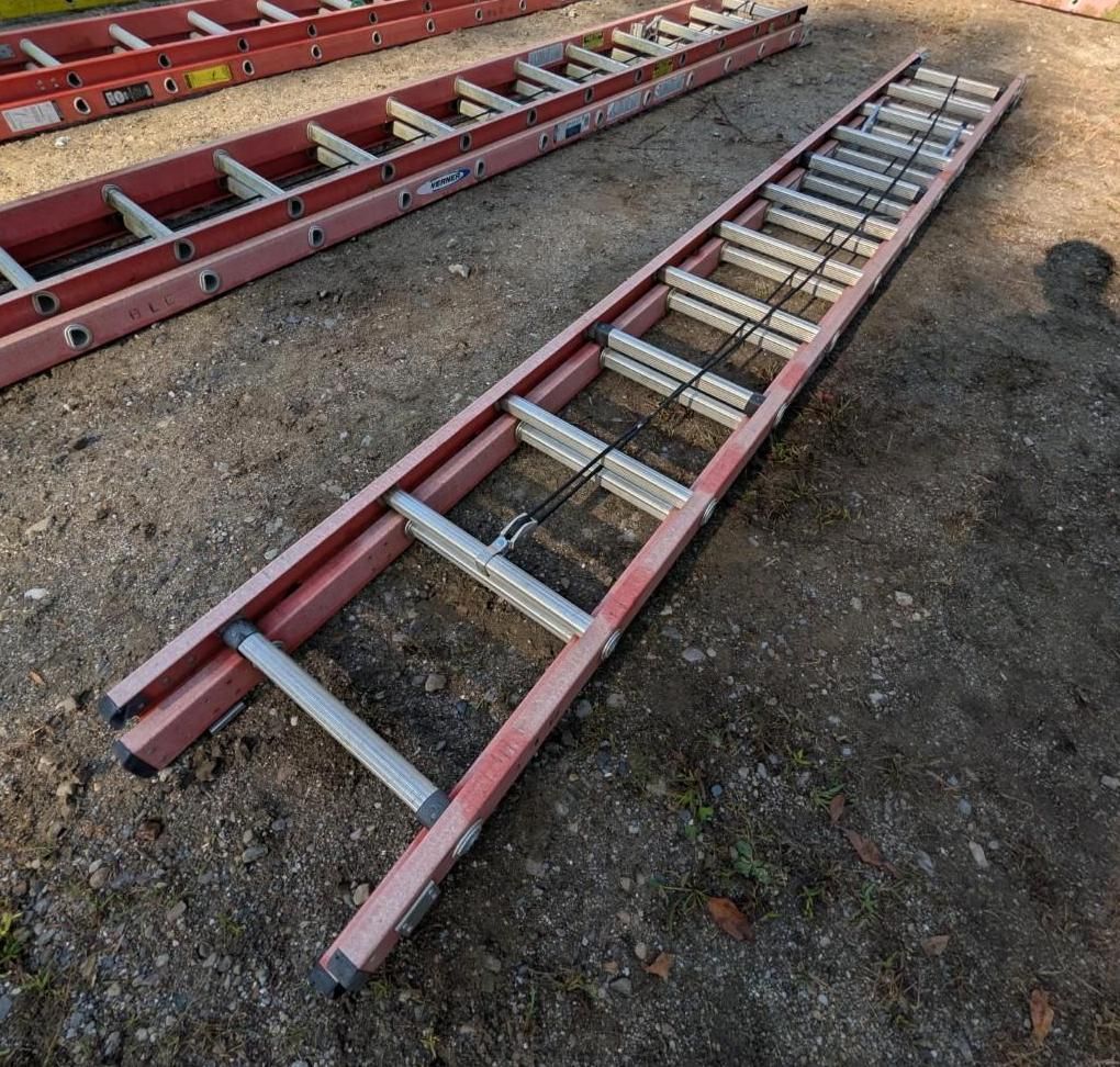 Red extension ladders lying on gravel outdoors, one stacked beside another.