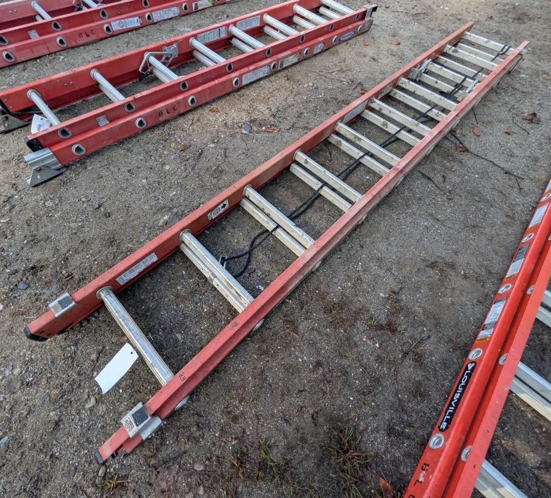 Red extension ladders stacked on gravel outdoors, with red rails and gray rungs.