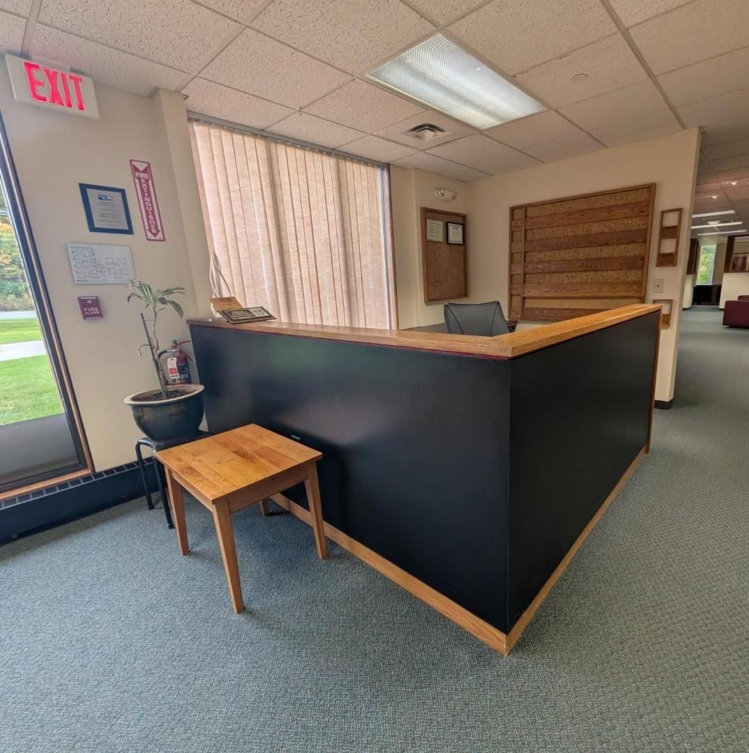 Quiet office lobby with a black reception desk, small wooden table, potted plant, and EXIT sign near windows.