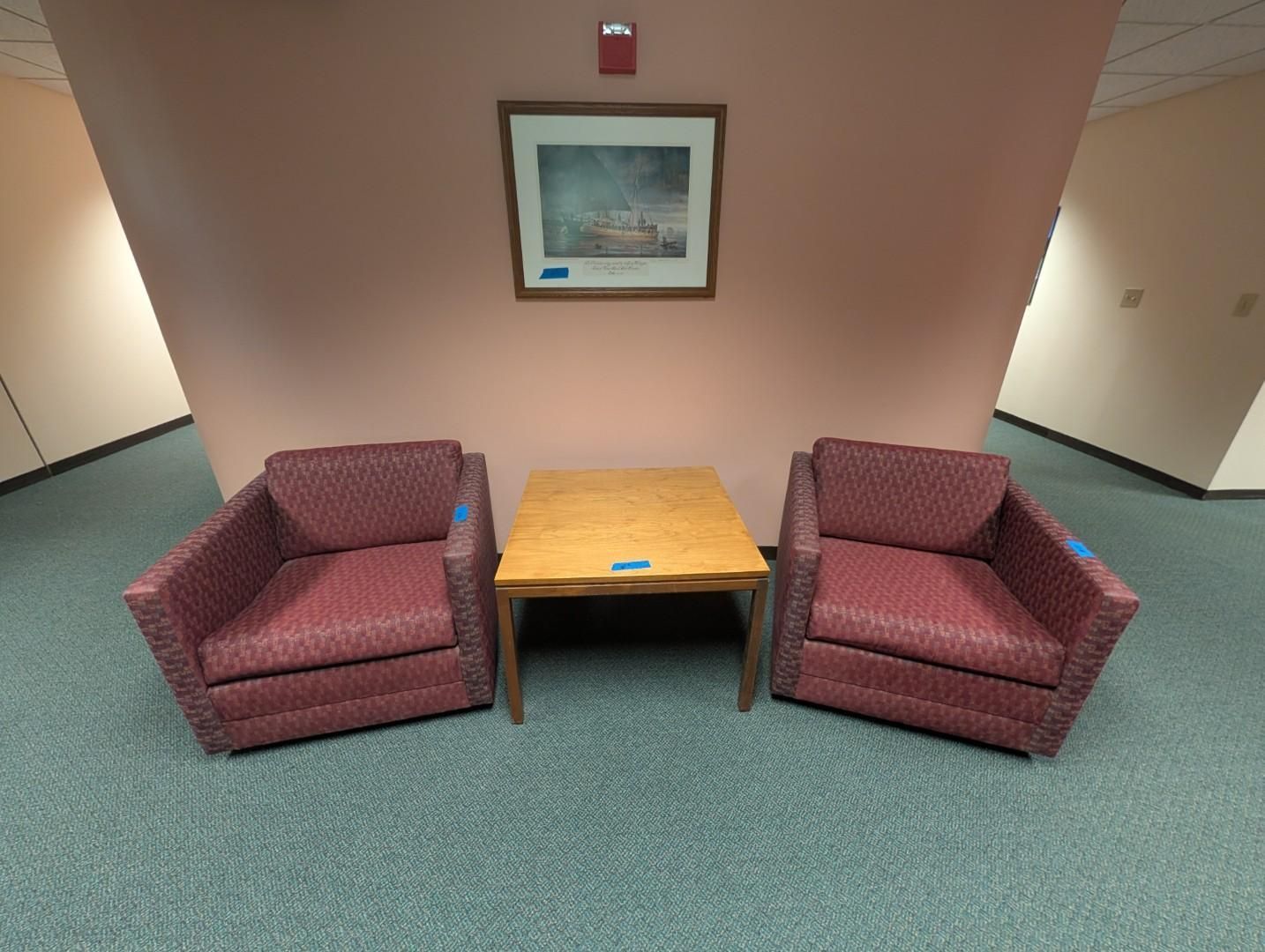 Two maroon armchairs flanking a wooden table in a hallway lounge area with a framed picture above.