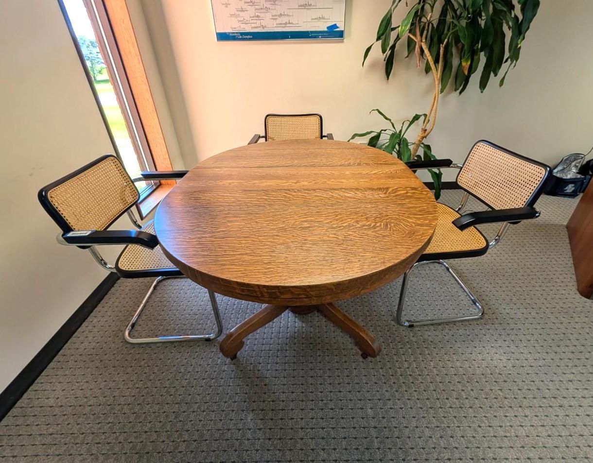 Round wooden conference table with four chairs in a carpeted office corner near a plant and window.