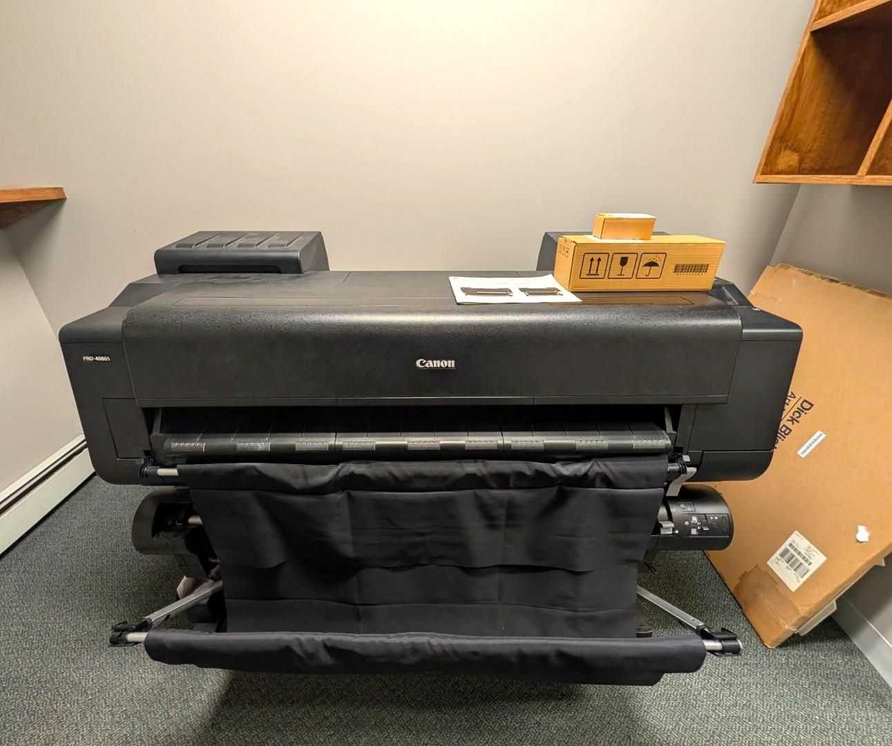 Large black Canon wide-format printer in a room, with paper roll and cardboard boxes nearby.