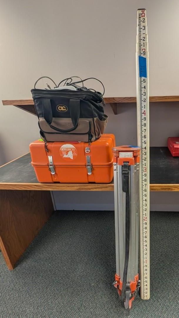 Stacked orange suitcases with a black bag and folded white walker beside a desk.