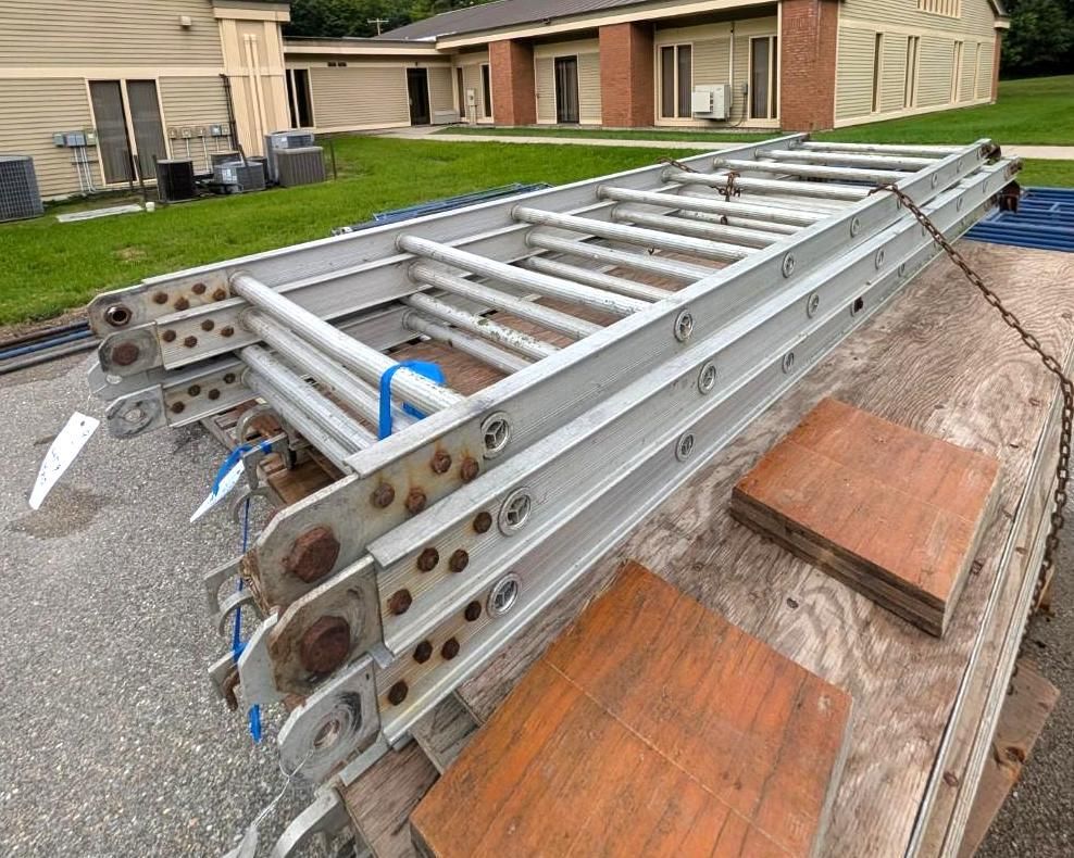 Stacked aluminum extension ladders on a flatbed trailer in a residential parking lot.
