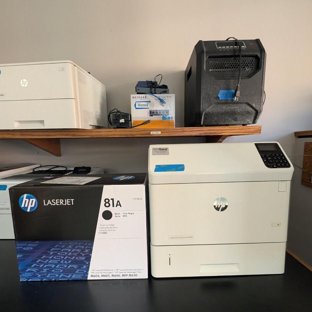Two HP printers and printer boxes stacked on a shelf in a storage area