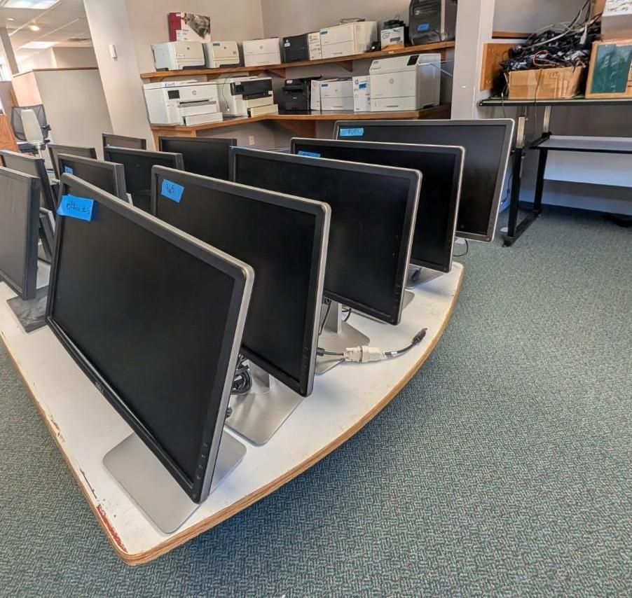 Row of black desktop monitors on a display table in a storage room with shelves and boxes