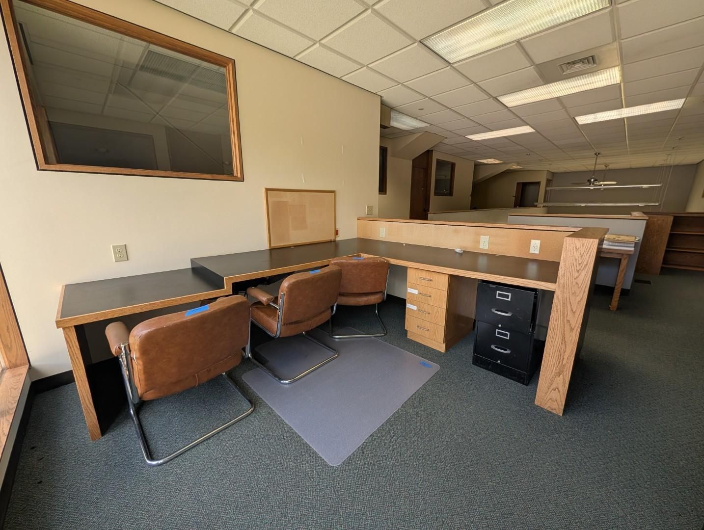 Empty office cubicle with desks, chairs, and filing drawers under fluorescent ceiling lights.
