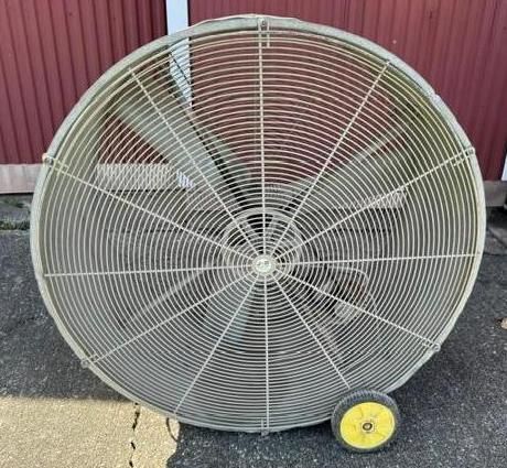 Large industrial floor fan with a metal grille and yellow wheels, standing outdoors on pavement.