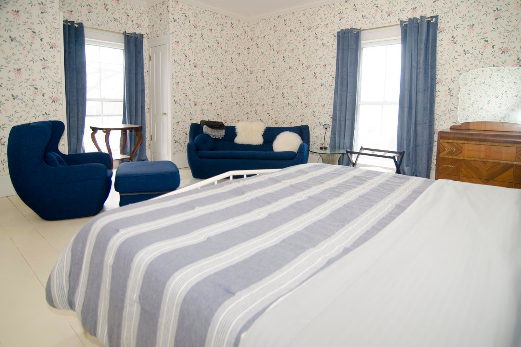 Bedroom with blue accents: striped blanket, loveseat, armchair, curtains, floral wallpaper, wooden dresser, window.