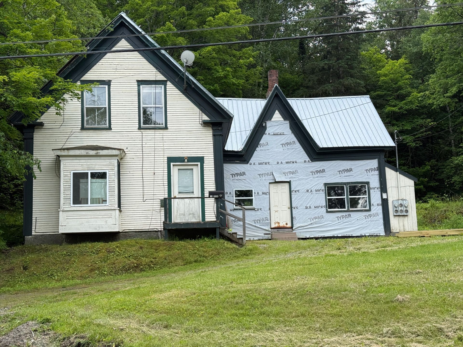 Two small white houses with dark trim on a grassy lawn, backed by trees
