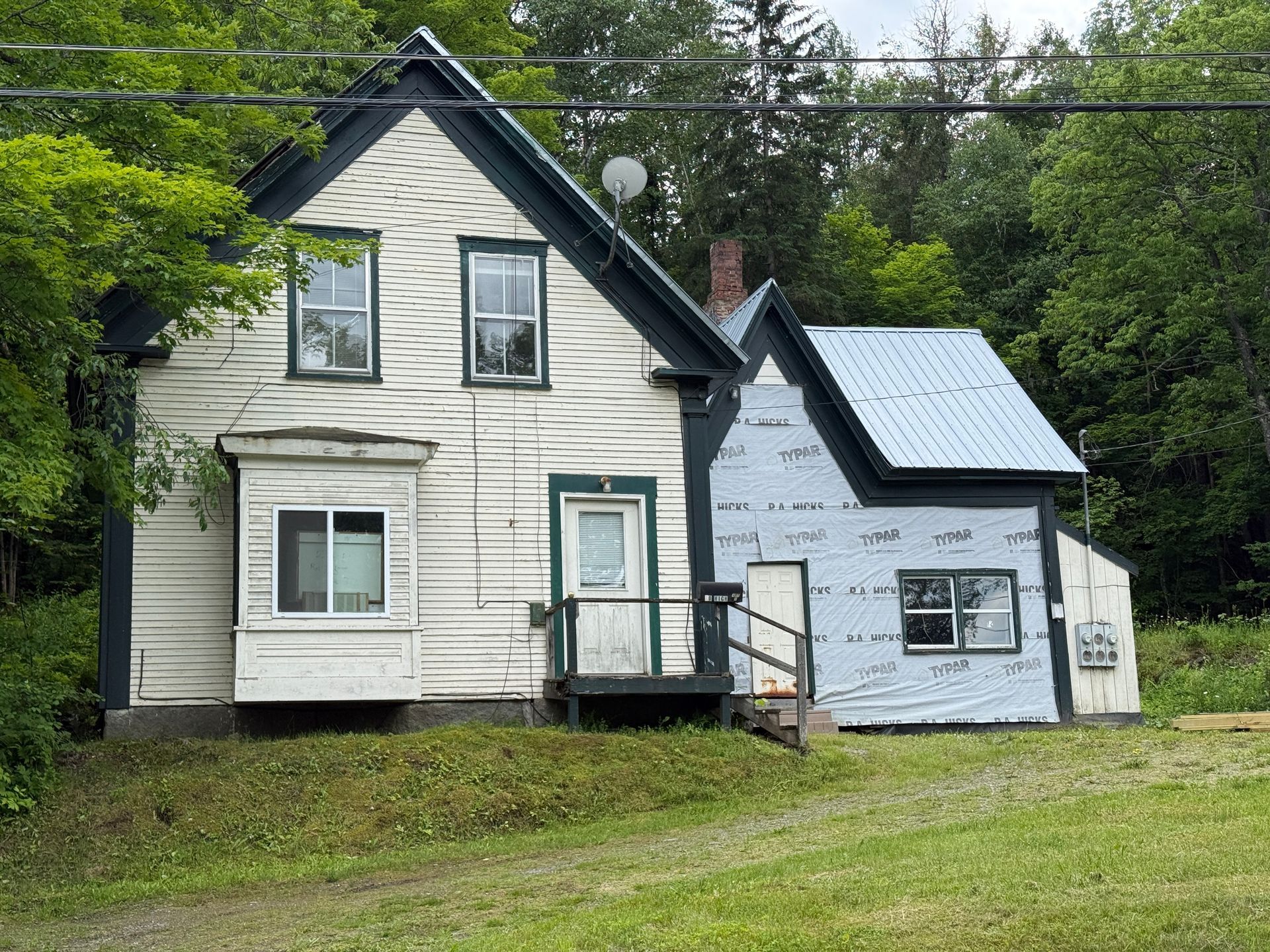 Small white house with dark trim and a light blue addition, set in a grassy wooded area.