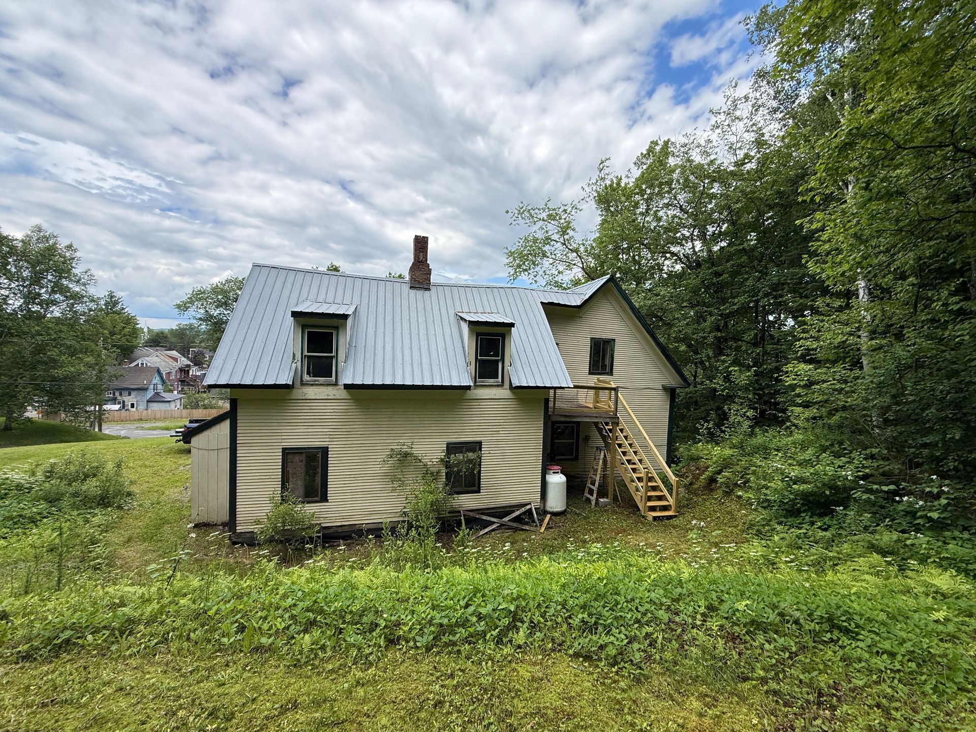yellow house with gray roof and porch stairs, surrounded by grass and trees under a cloudy sky