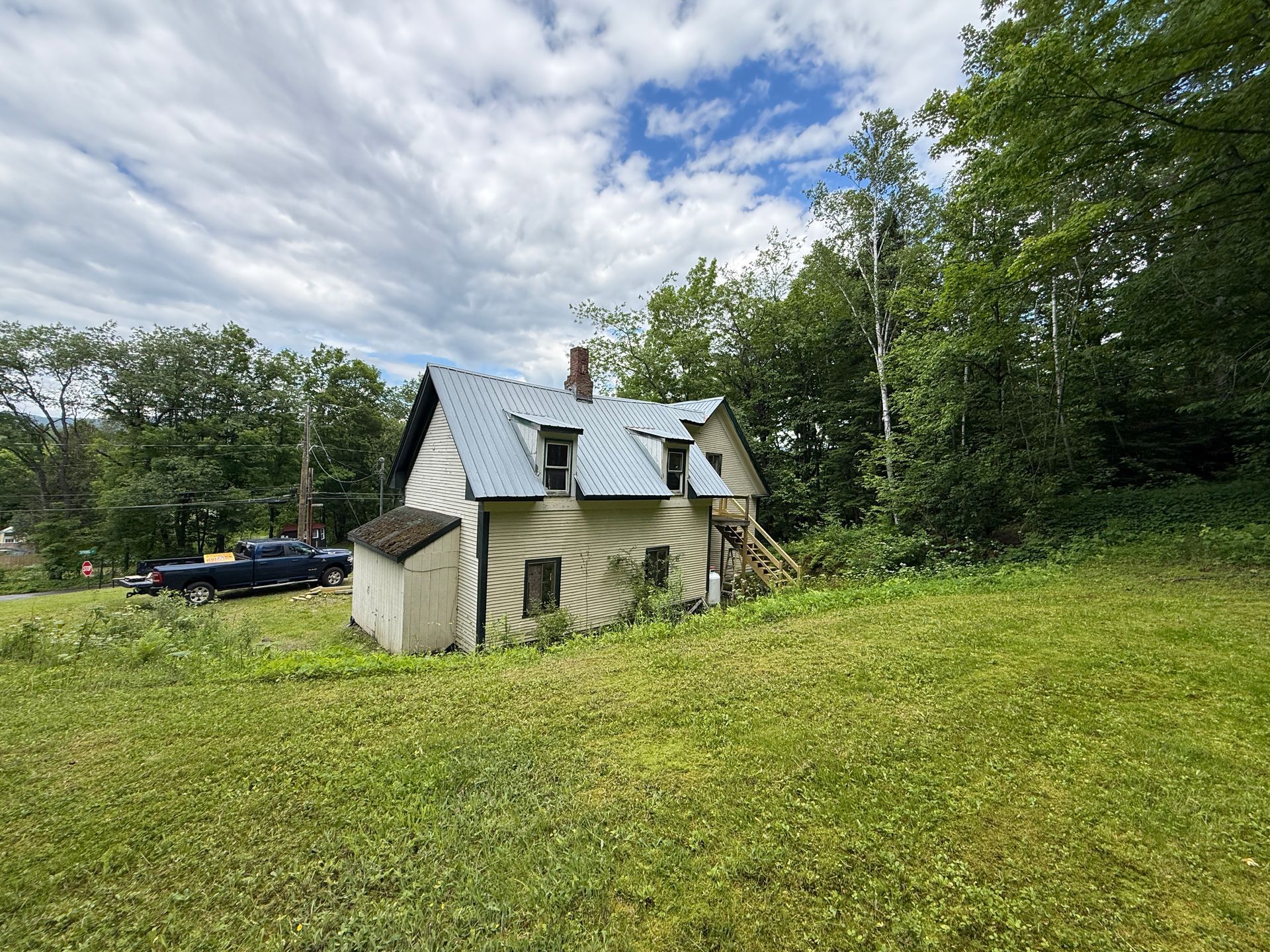 Small white cottage with metal roof in a grassy field, surrounded by trees under a cloudy sky