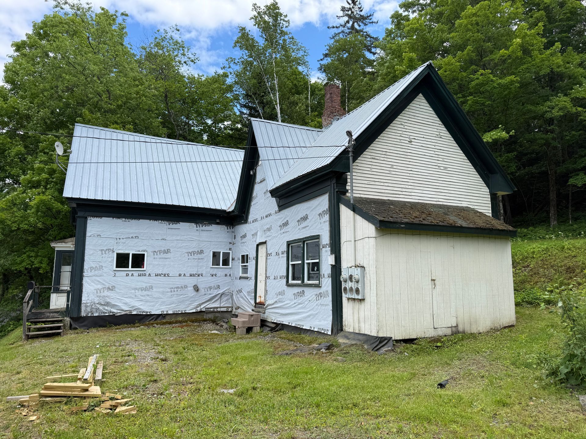 Old white house with a gray metal roof on a grassy hillside, surrounded by trees under a blue sky