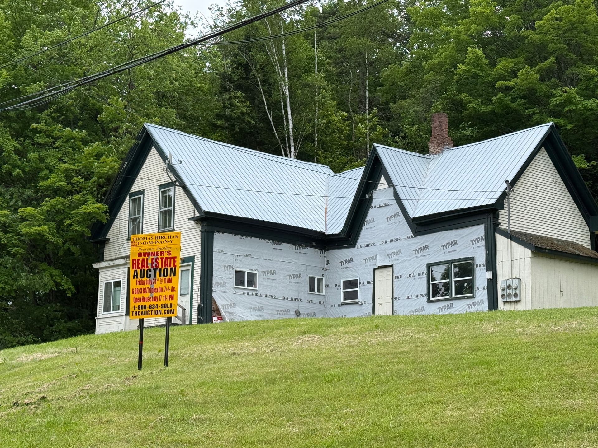 White house with black trim on a grassy hillside, with a bright yellow for-sale sign in front.