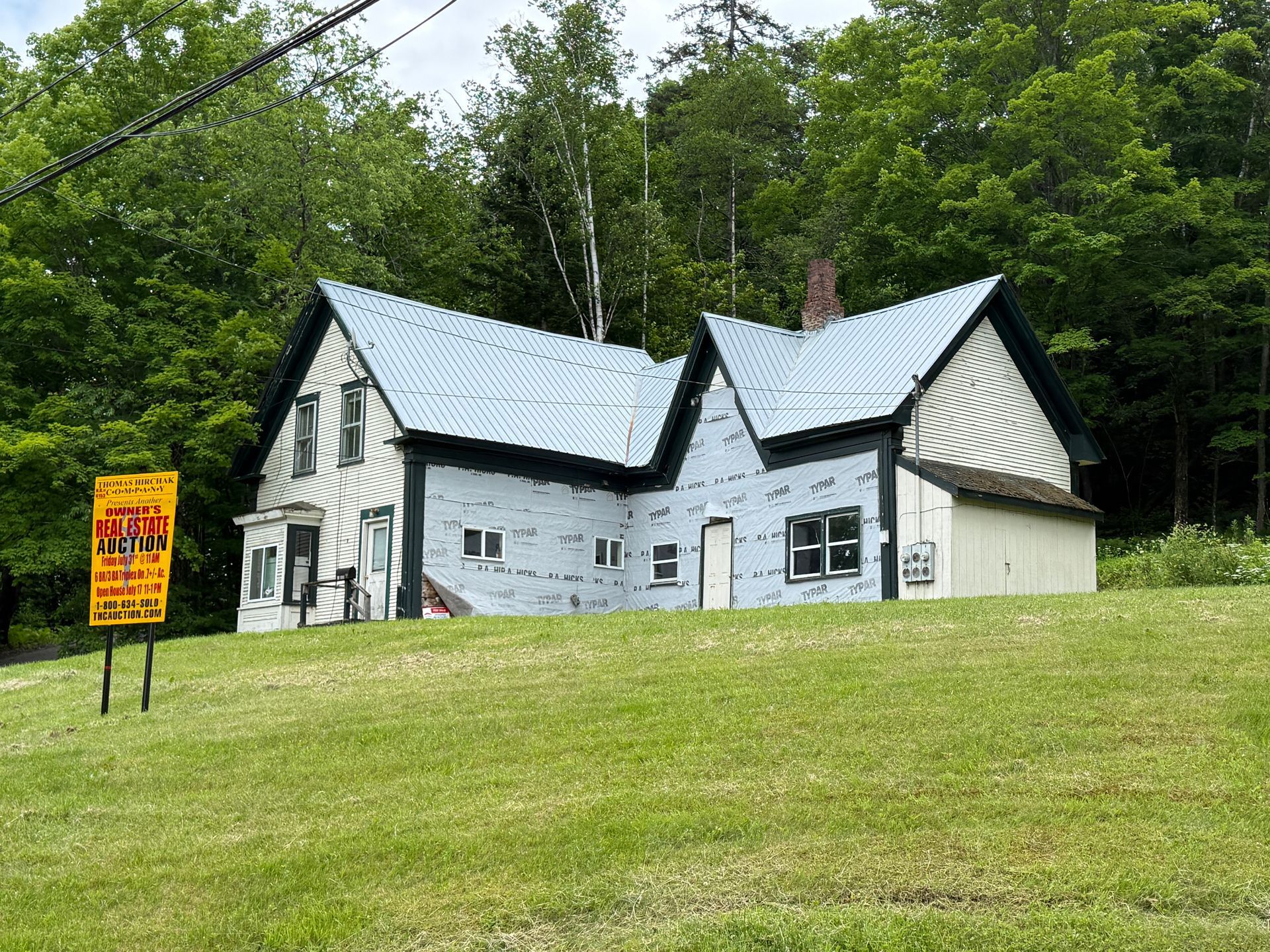 White house with black-trim roof on a grassy hill, surrounded by trees, with a small sign in front.