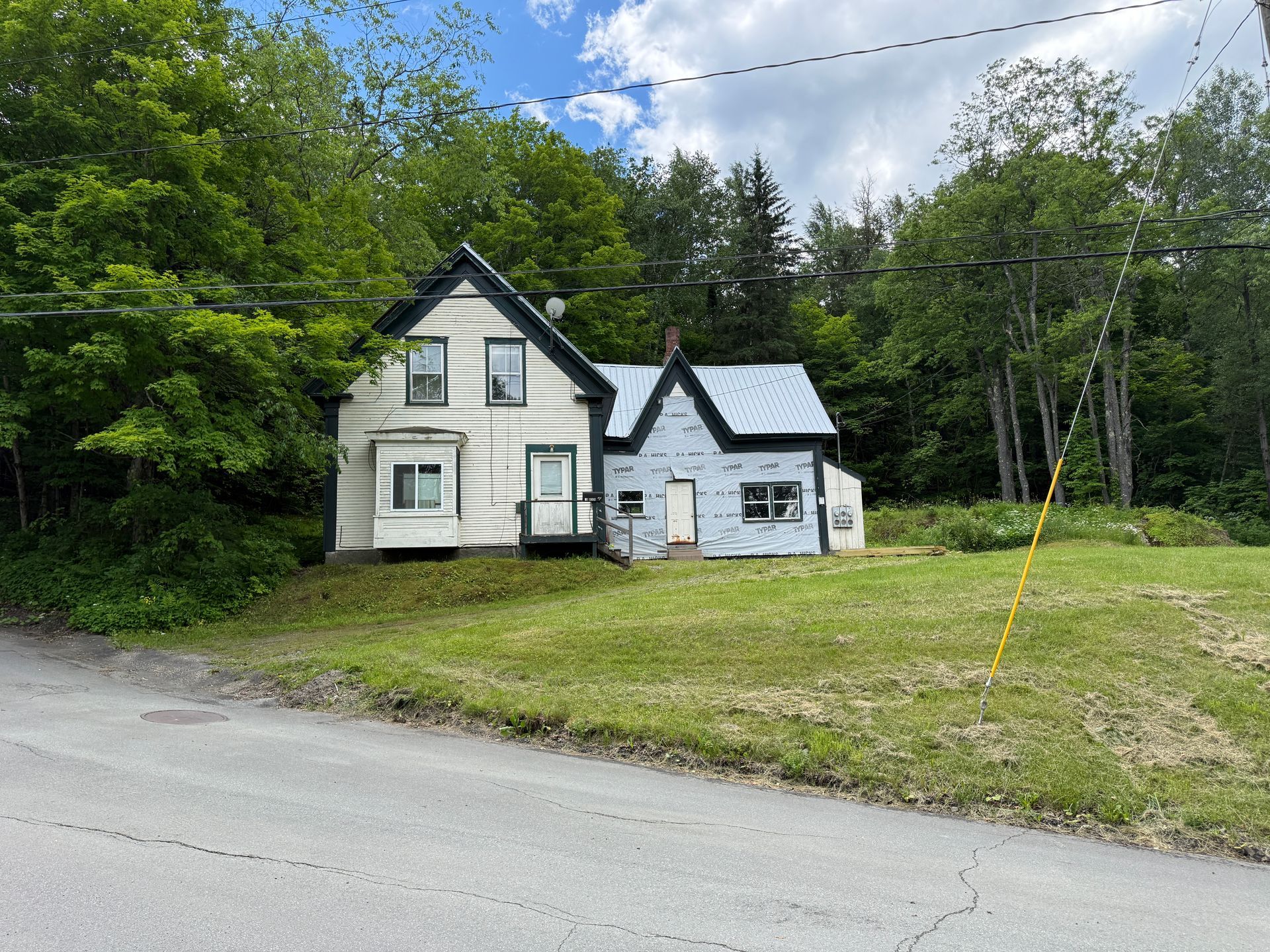 White house and small outbuilding beside a road, surrounded by trees and grass under a cloudy sky