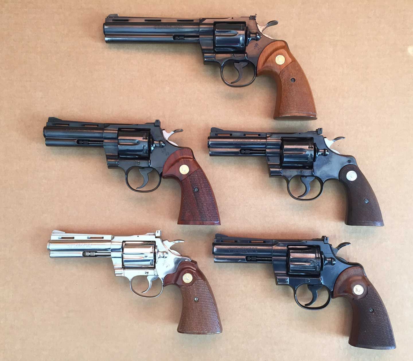 Five Colt Python revolvers, blued and stainless, with wood grips, on a light brown surface.