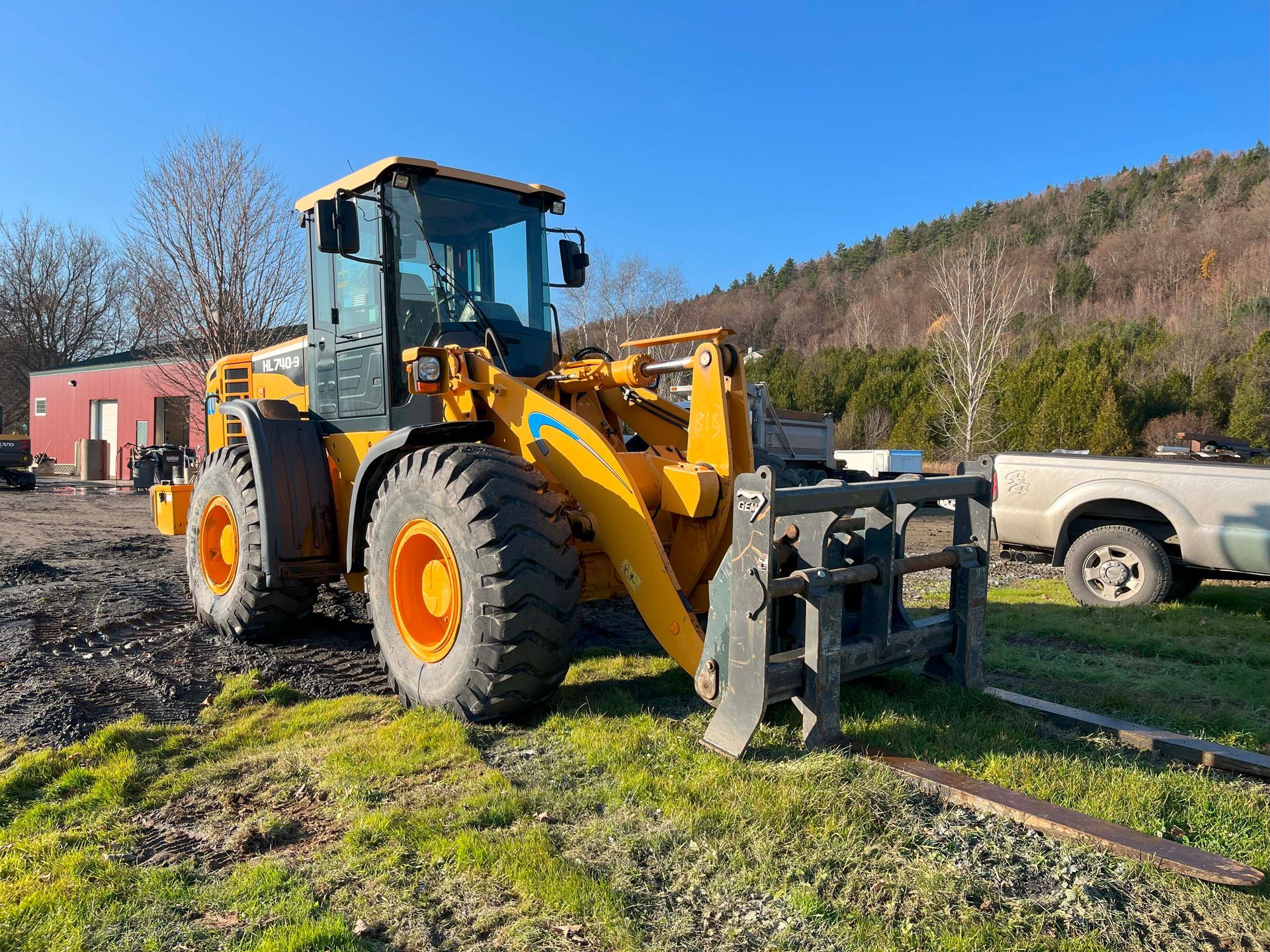 Yellow front loader on a grassy dirt lot beside a pickup truck, with a red container and hills in the background