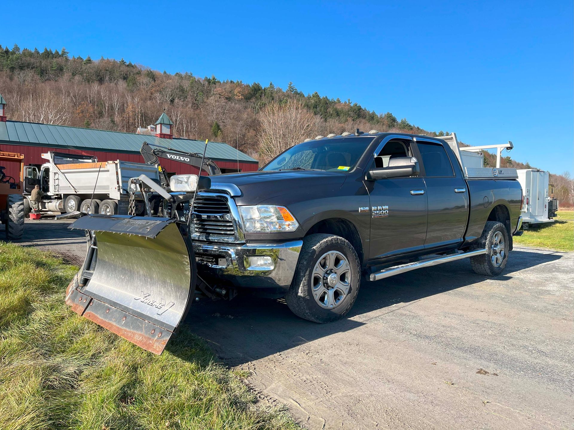 Black pickup truck with front plow parked outdoors on a gravel lot with buildings and trees in the background