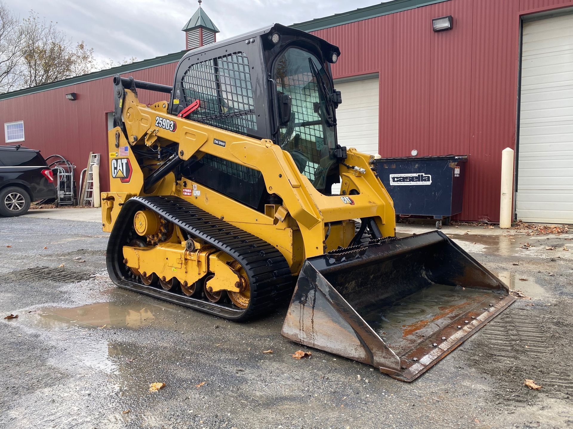 Yellow tracked skid steer loader with front bucket parked on wet gravel beside a red building.