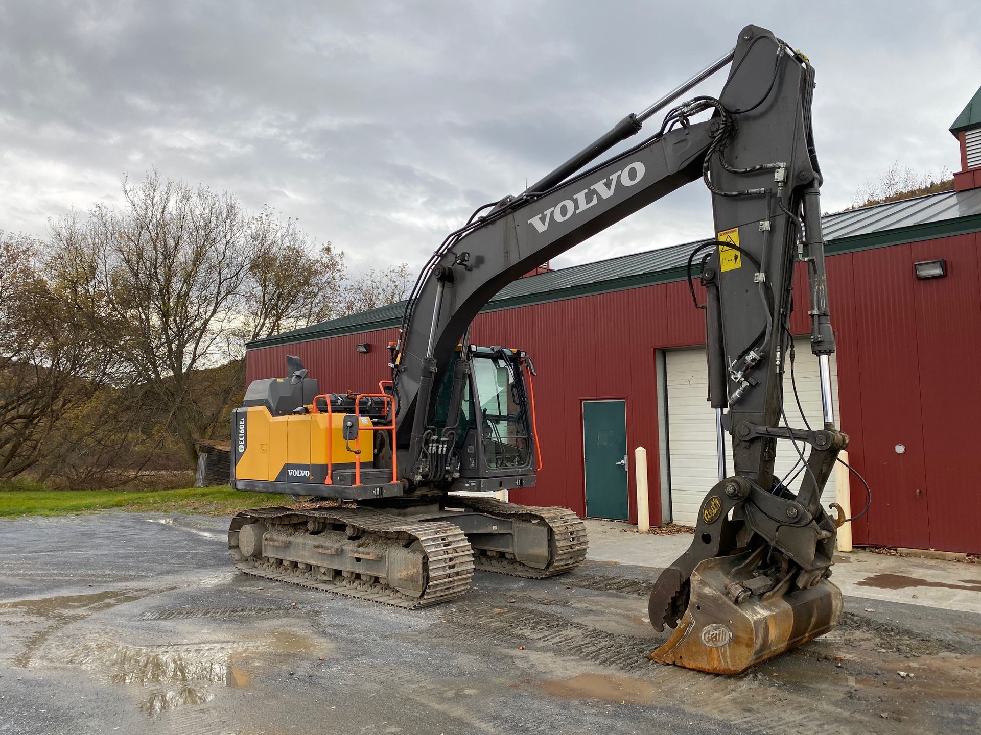 Yellow Volvo excavator on ground beside a red building on an overcast day