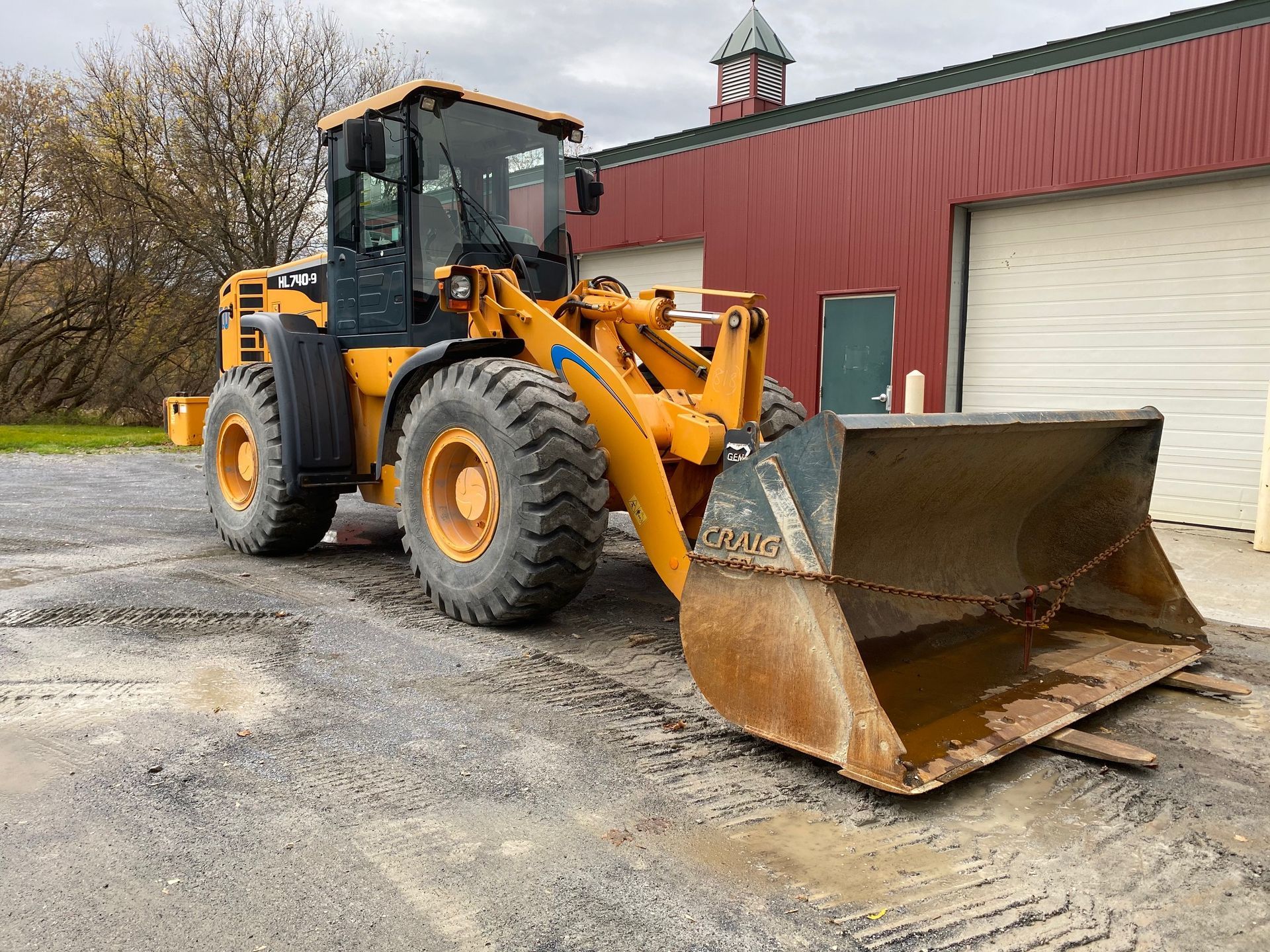 Orange front loader parked beside a red barn on a wet gravel lot, bucket lowered.