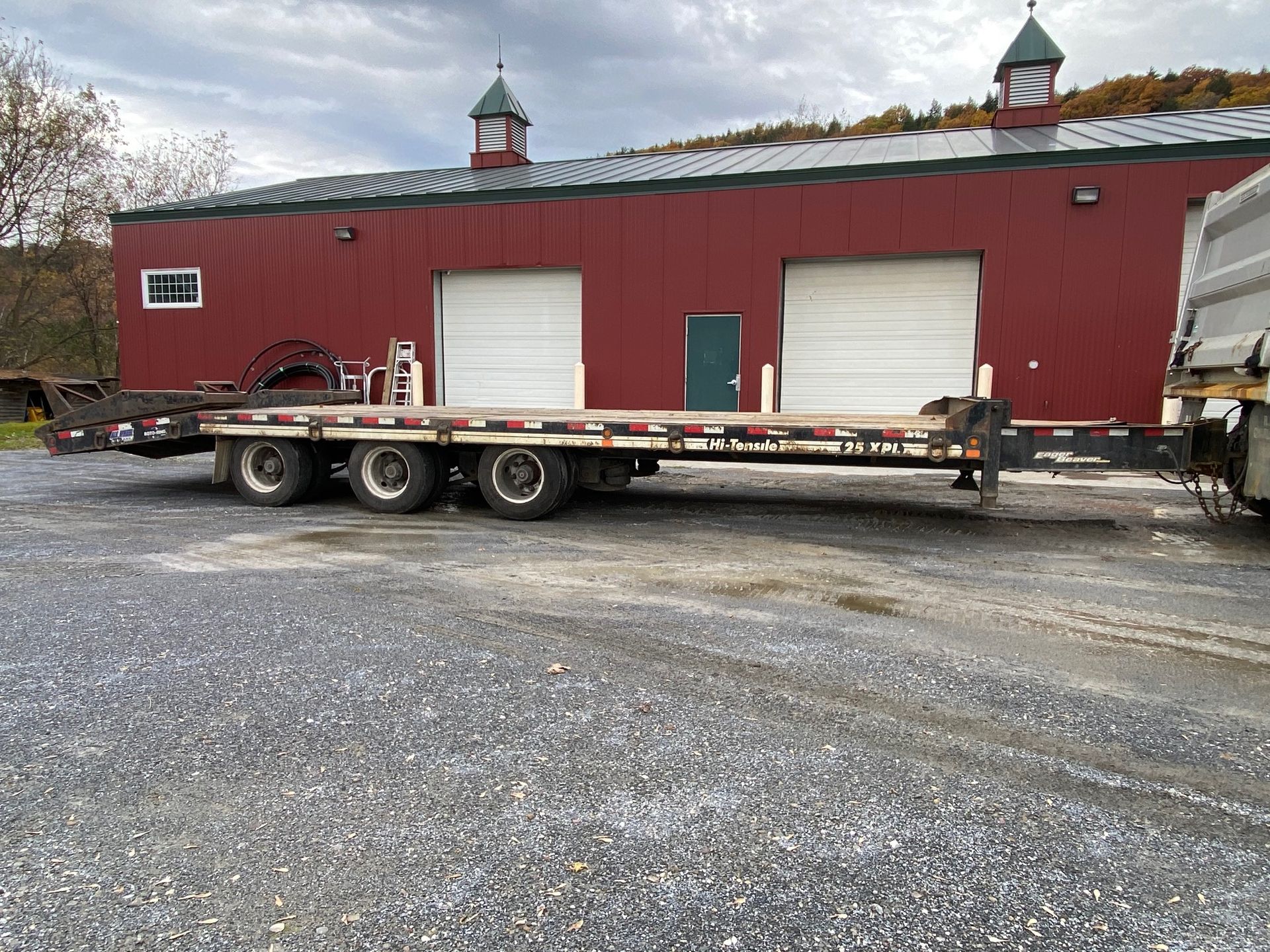 Flatbed trailer parked beside a red barn with white garage doors on a gravel lot