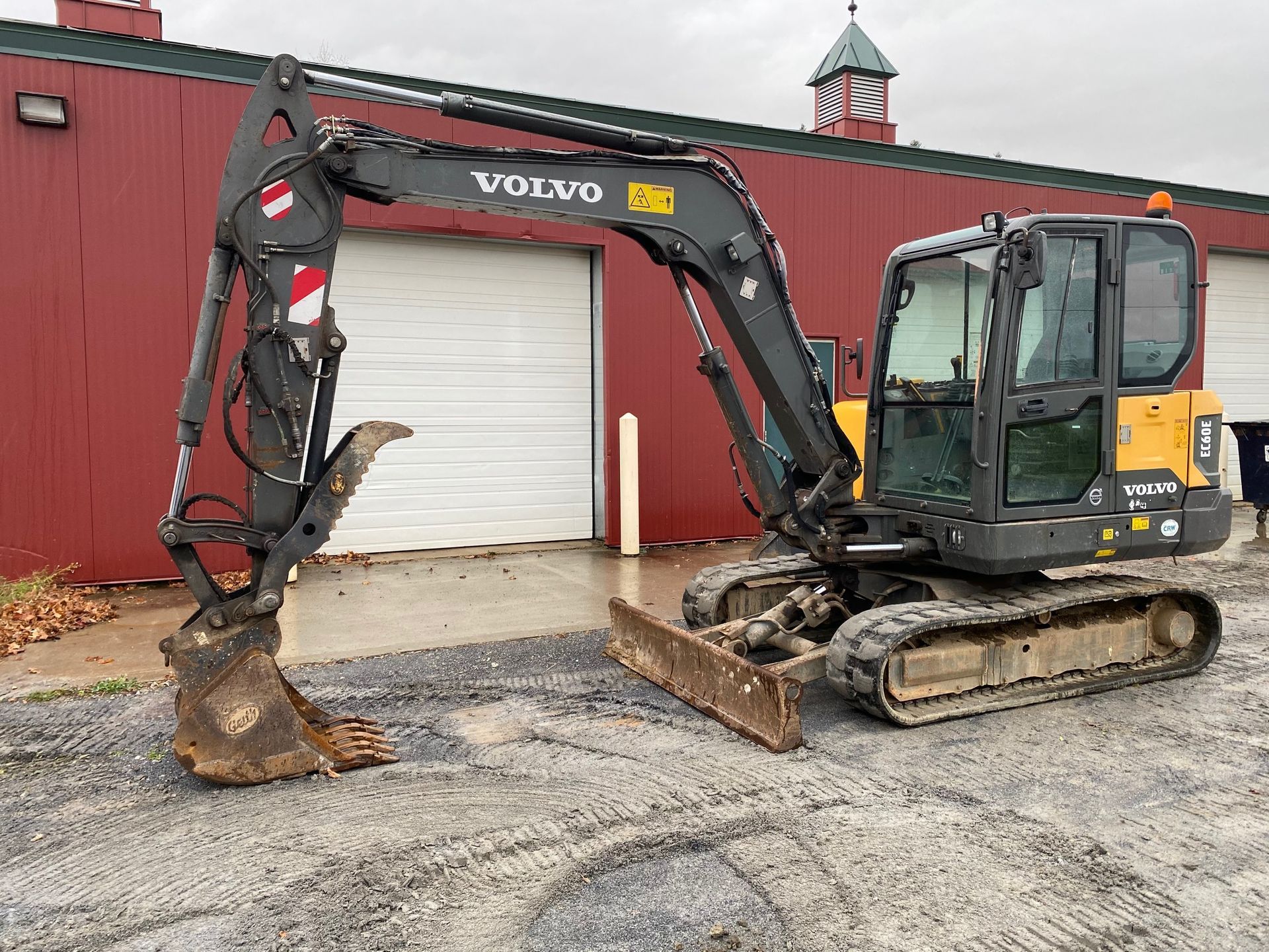 Yellow Volvo excavator parked beside a red building with a white garage door