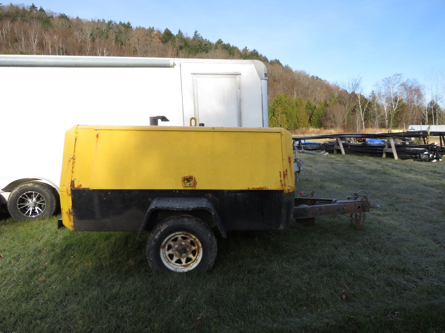 Yellow-and-black trailer parked on grass beside a white truck, with wooded hills in the background