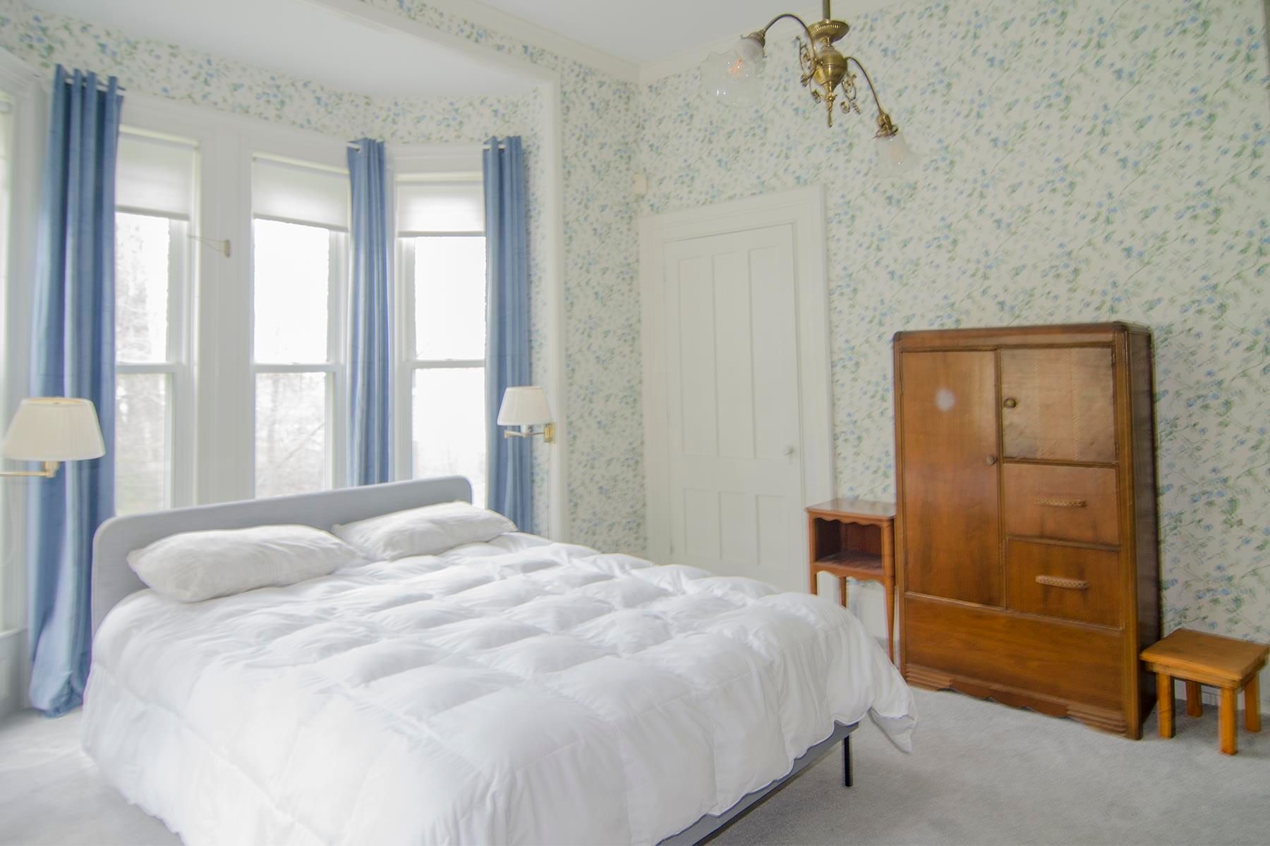 Bedroom with white bedding, blue curtains, patterned wallpaper, and wooden furniture.