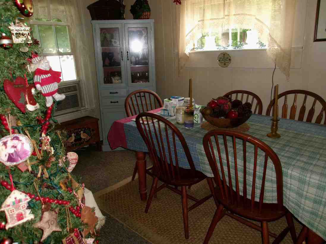 Dining room with a decorated Christmas tree, table set with a blue cloth, and wooden chairs by sunlit windows.