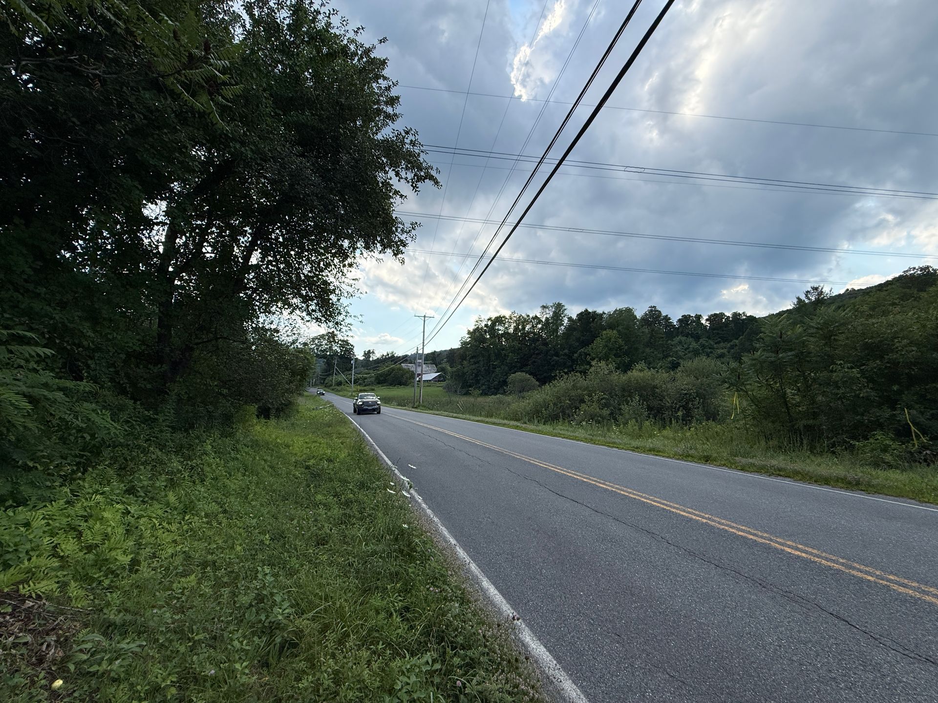 Rural road with a car in the distance, trees on both sides, and power lines under a cloudy sky