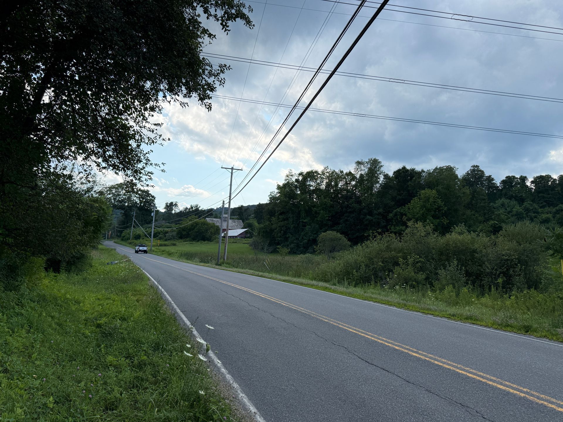 Quiet rural road curving past trees and grass under a cloudy sky, with power lines overhead