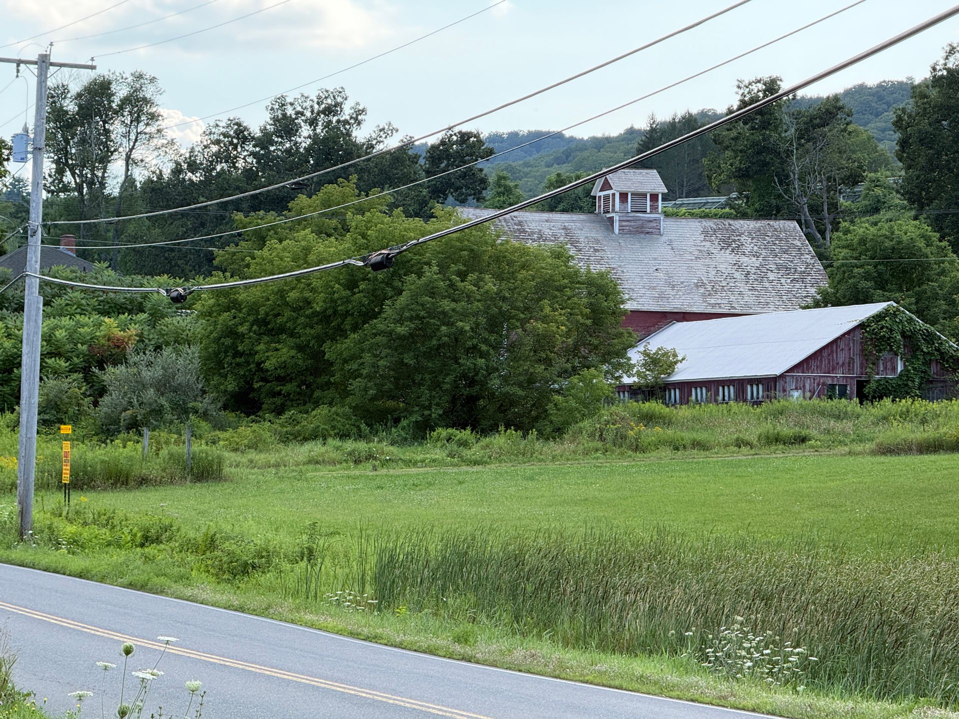 Rural roadside with green fields, a red-roofed barn, trees, and power lines under a cloudy sky