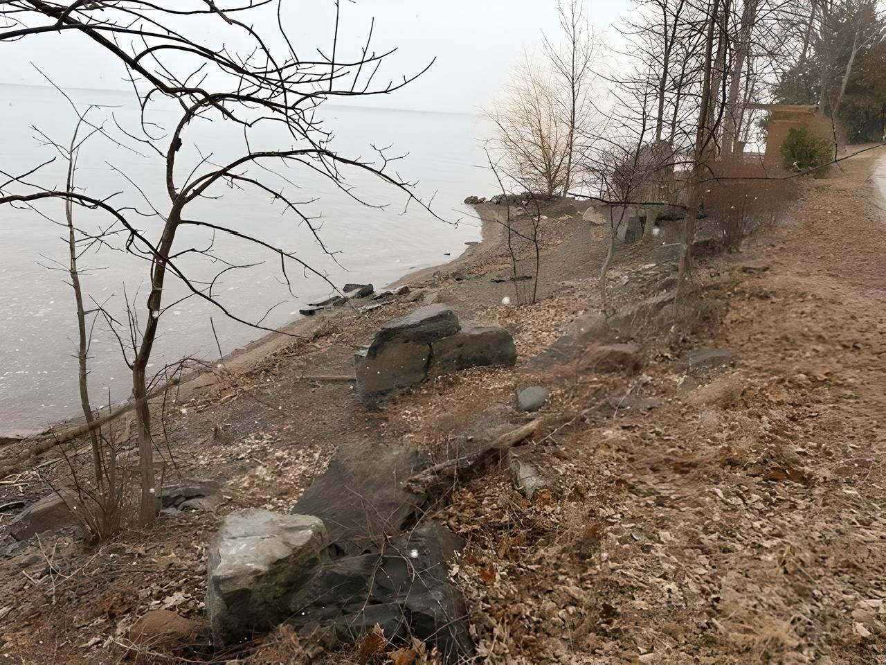 Shoreline next to a path with trees, gray water, and scattered rocks.