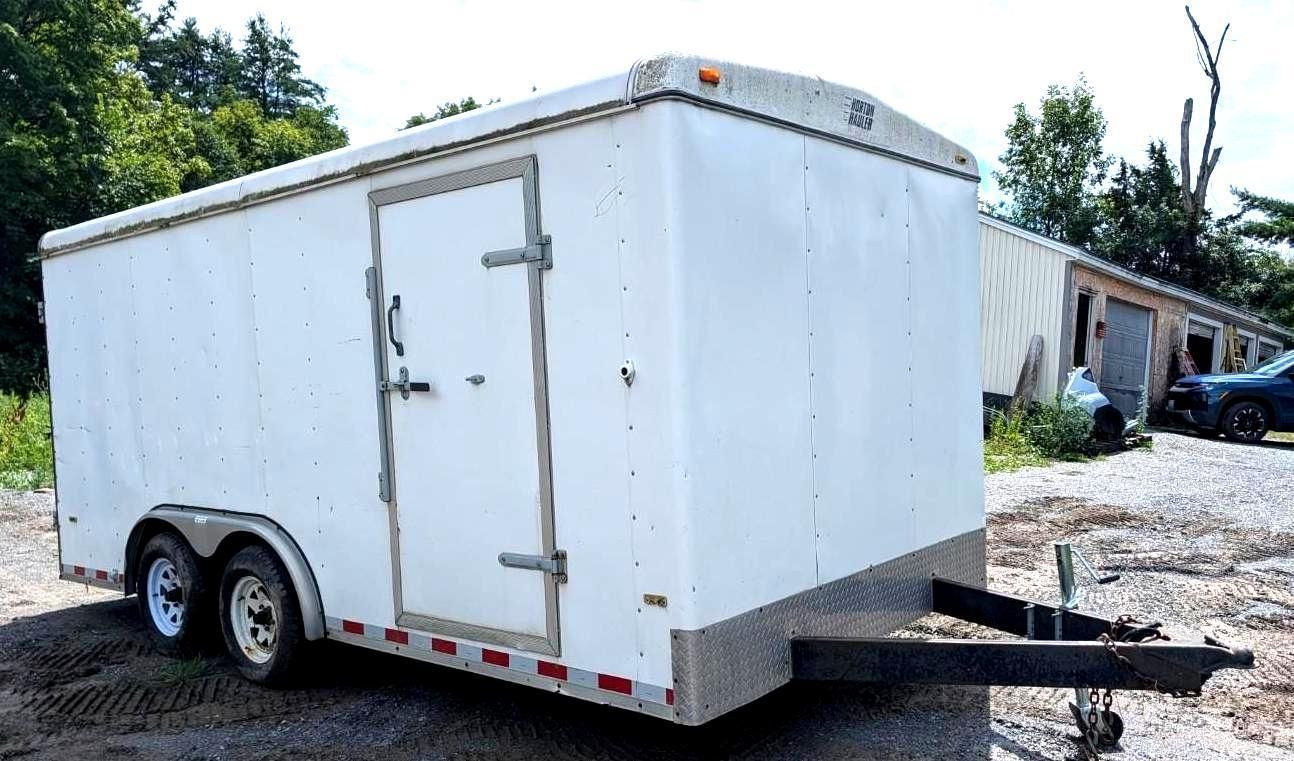 White enclosed dual-axle cargo trailer parked on gravel beside a building, with tow hitch visible.