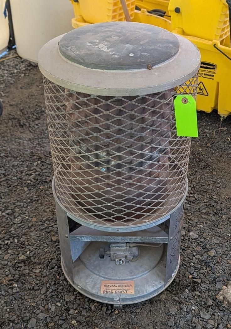 Gray cylindrical metal wire-mesh heater/stove with a dark top and green tag, outdoors on gravel.