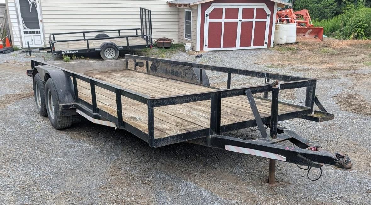 Black utility trailer with ramps parked on a gravel driveway in front of a garage and yard