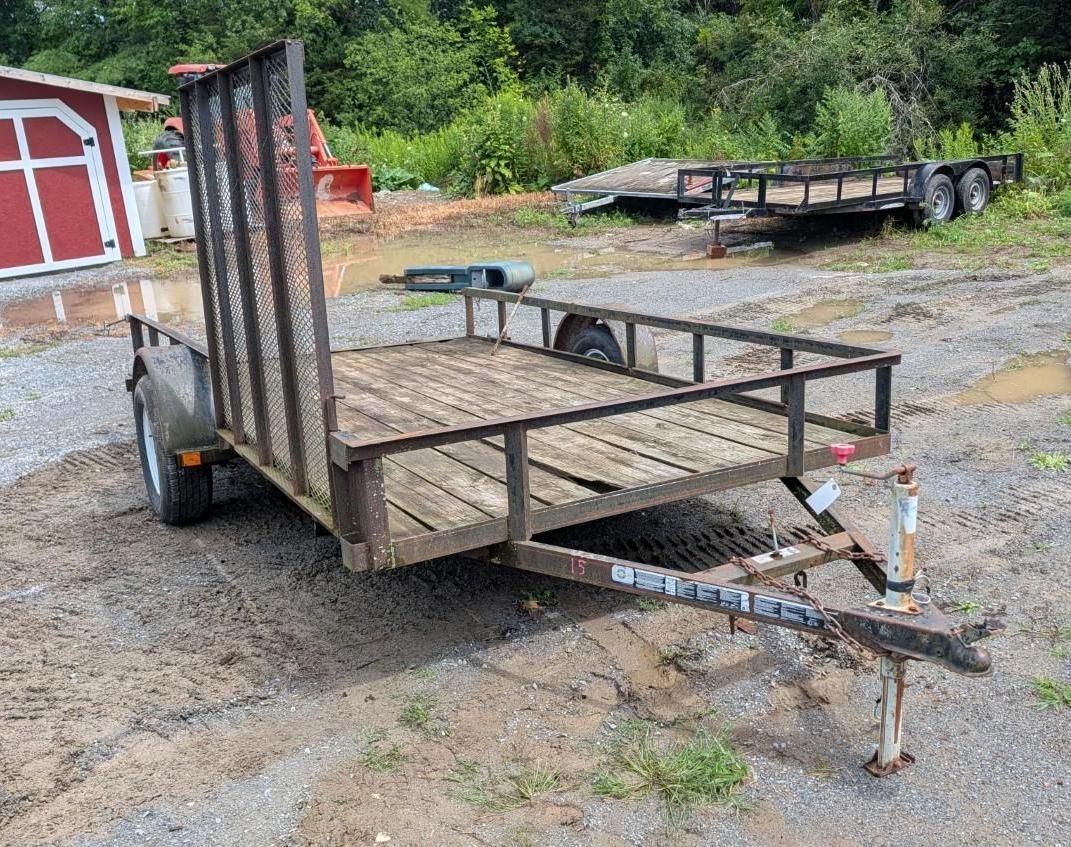 flatbed utility trailer on gravel near a red shed and trees.